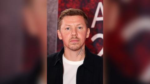 Professor Green has short blonde hair and is wearing a white t-shirt under a black jacket