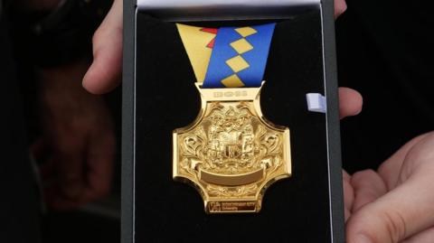 A highly-detailed gold cross-shaped medal on a multi-coloured ribbon in a black case