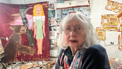 Rose Wylie on the right, facing the camera, in glasses, dark top and multi-coloured scarf. Behind her on the left is a painting of a blonde-haired woman, a hammer and a bear.