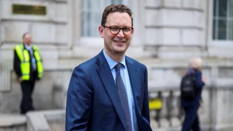 Darren Jones, who has short brown hair and glasses, smiles at the camera as he stands in front of the stone-fronted building of Whitehall. He is wearing a medium blue suit, a blue shirt and a dark patterned tie.