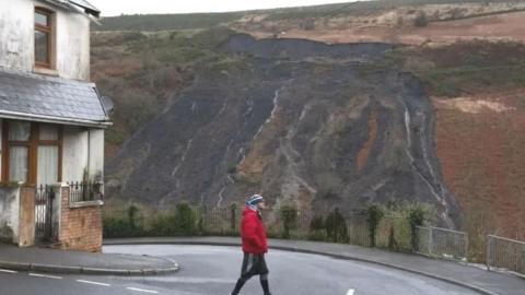 A man in red top and black leggings walks across a road with the damage from an old tip sliding down the mountain in the background