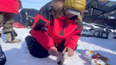 A man in kneeling on the ice in a red parka cutting a chunk of meat.