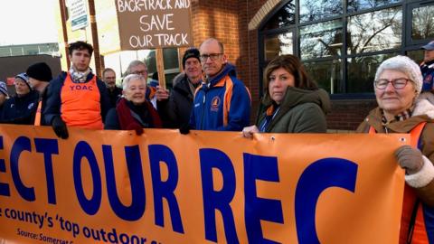 A group of protestors holding an orange and blue banner reading 'protect our rec'