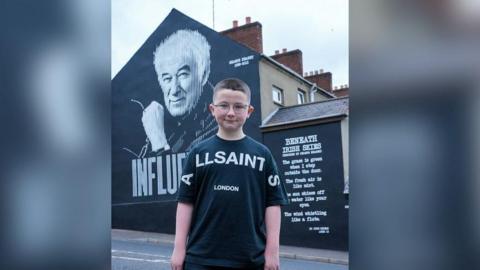A boy , who is dressed in black t shirt and trousers, stands in front of a gable wall on which a mural in memory of Seamus Heaney is painted.