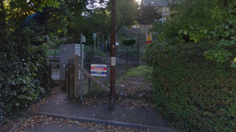 A gated pedestrian railway crossing surrounded by trees and hedges. A warning sign and railway information board are near the entrance, with tracks running through a wooded area.