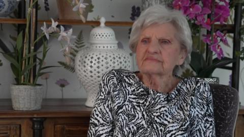 A woman with grey wears a black and white top and sits in front of an oak dresser with an orchid and vase on it.