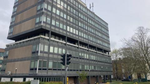 A grey and glass seven-storey office block with traffic lights and some bare trees outside
