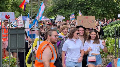 A large crowd of people walking through the metal gates of an outdoor park during the Pride event in Bath. Many people are holding up pride flags or handmade signs expressing support for LGBTQ+ rights.