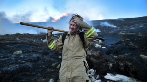 Hayley Agnew standing with her hand held to her forehead, and her shovel held across her shoulder. Her face is scrunched up in distress, and smoke and fire is visible behind her