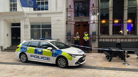 A police car is parked partly on a kerb outside a building in a street. There are dark benches outside, with blue and white police tape in front of them. Two officers are standing outside brown doors. 