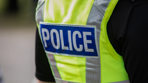 The back of a police officer's uniform, showing a hi-vis green jacket with silver stripes, saying 'police' in a blue patch on the jacket