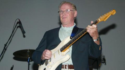 Vic Flick performing on stage. He is wearing glasses, a suit jacket and playing a white electric guitar
