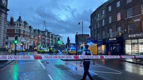 A red and white police cordon at the scene in Clapham, with several police cars and an ambulance in the distance