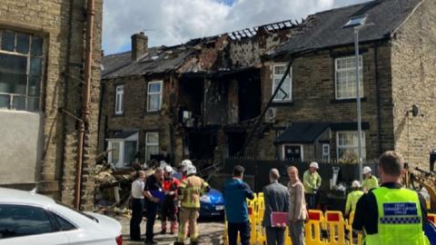 Emergency services crews and other stand around a cordoned off street. In the centre of the frame is a terraced house with much of its upstairs missing.