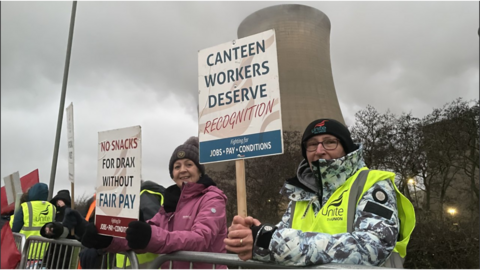 Workers stand on the picket line outside Drax power station. They're are wearing high-vis and holding placards. A cooling tower can be seen in the background.