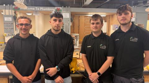 Four young men standing next to each other in a workshop with two on the left wearing black hoodies and the other two wearing black polo shirts and a sign saying 'Welcome to Plumbing' on the wall behind them.