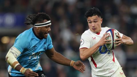 Elia Canakaivata (left) playing for Fiji, wearing a white headband, attempts to tackle England's Marcus Smith who is carrying the rugby ball