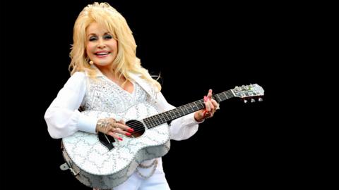 Dolly Parton dressed in white with a guitar on a black background. 