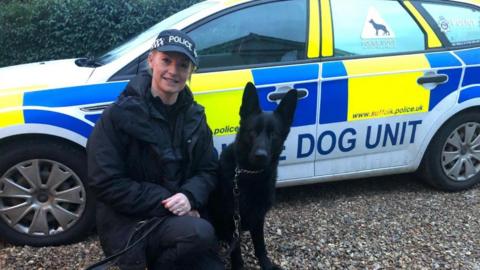 A photograph of a female police officer kneeling next to a black German Shepherd dog. There is a "Dog Unit" police car in the background.