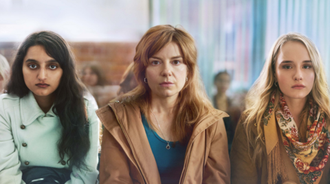 This image shows three women sat side by side, the woman on the left hand side has long brown hair, brown eyes and is wearing a light blue coat. The woman in the middle has long ginger hair, brown eyes and is wearing a dark blue top and brown jacket. The woman on the right hand side has blonde hair, brown eyes, and is wearing a yellow floral patterned scarf and a brown jacket. All the woman have blank, emotionless expressions. 