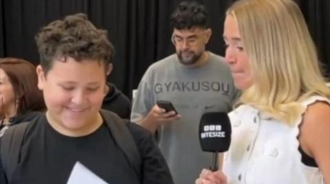 Student in a black tshirt smiles and looks at results paper. He stands next to BBC presenter