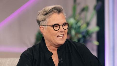 Rosie O'Donnell sits on a sofa and appears to be in a TV interview. She has short grey hair and is wearing black round glasses and a black shirt.