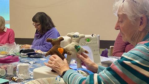 Jean Sampson is sitting at table holding two sensory bears near three other women who are sewing. In front of Mrs Sampson is a sewing machine and there are reels of thread, swathes of fabric and sewing tools spread across the table.