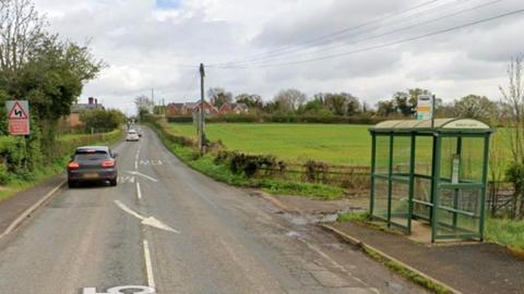 A rural road which has a green field on one side of a road. Next to the field is a bus shelter. There are two cars on the road and arrows and road markings. 