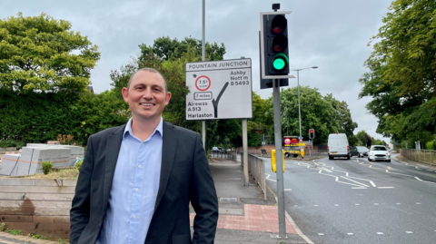 A man in a dark blazer and blue shirt is smiling, and stands in front of a road sign detailing a junction. The road on which the sign stands is lined by trees.