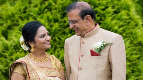 A man and a women stand outdoors against a backdrop of green leaves, wearing detailed traditional outfits. They are standing close together and looking away from the camera and at each other, smiling.