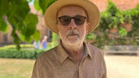 Philip Roberts, a man wearing sunglasses as well as a peach-coloured shirt and a straw hat, is stood in front of an English Tudor building. There is a lawn behind him and a series of low hedges.