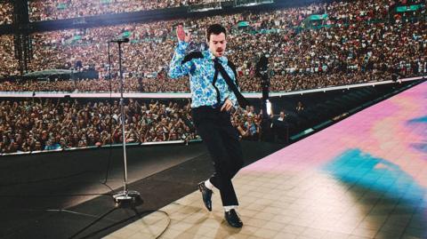 Harry Styles dancing on stage at Wembley wearing a blue floral shirt, black tie, black trousers, white socks and black patent shoes. Thousands of people can be seen standing or watching him from the stands