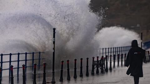 A woman stands on a seaside promenade as waves crash over