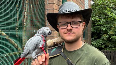 A picure of Connor Atherton who is wearing a hat and green t-shirt and black glasses. He smiles into the camera, whilst holding a gray parrot.