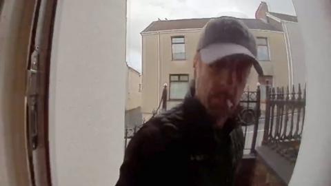 Richard Jones seen in doorbell camera at front door wearing a cap and smoking a cigarette