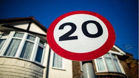 A close-up of a 20mph circular road sign, with a row of houses behind.