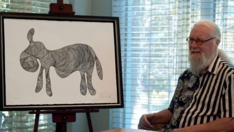 Sir Billy Connolly sitting beside a framed picture of his drawing "Drunk Donkey"