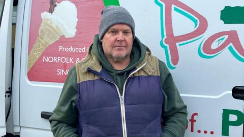 Adrian Nichols wearing a grey woollen hat and a blue coat stands in front of a white van displaying a picture of an ice cream cone.