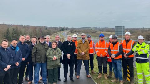 A large group of people standing on the side of a road. Some are wearing orange and yellow high vis suits and other members of the group are in long coats.
