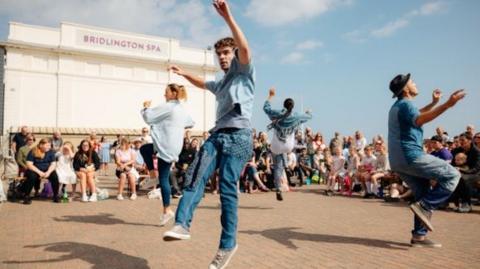 Four street performers, made up of two men and two women wearing denim jeans and denim trousers, are performing a dance on the street outside a theatre with people sat down watching them.