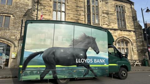 A Lloyds mobile banking van with an image of a black horse galloping on it parked on a road