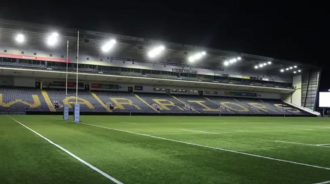 A side of a stadium reading "Warriors" across the seats, with a grass pitch below.