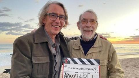 Steve Coogan (left) on set alongside Tom Michell. They are on a beach holding a film clapper-board for the movie The Penguin Lessons