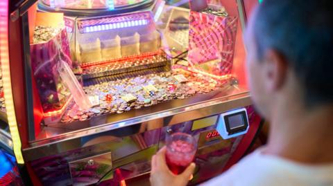 A man stares at an arcade machine with a drink in his hand. 