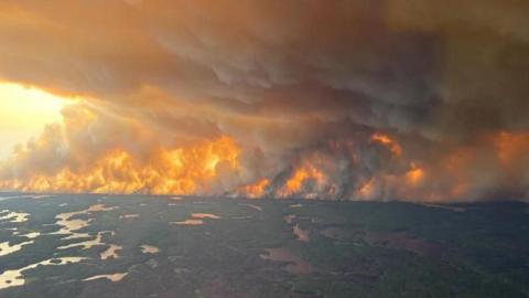 Image taken from a plane. You can see a wildfire on the horizon, with flames and smoke covering the landscape