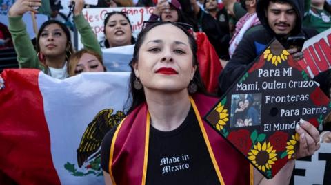 A woman looks directly into the camera with protesters behind her holding Mexican flags.