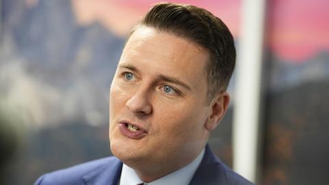Health Secretary Wes Streeting talking during a media interview. He has short dark hair and wears a navy suit with a white shirt and red tie underneath.
