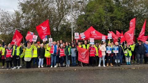 A large group of factory workers standing on a path outside wearing yellow or red hi-vis jackets. Many are holding red banners or flags branded with Unite the Union's logo. Others are holding banners that read 'pay up now!' There are trees behind them with green leaves and the sky is cloudy. 