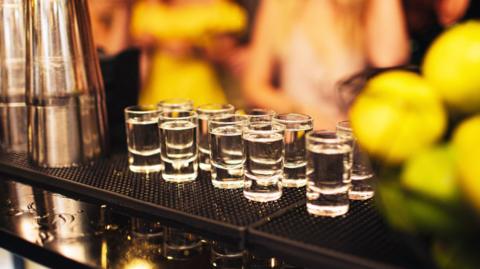 Ten shot glasses filled with clear liquid lined up on a bar next to a cocktail shaker. There are people blurred in the background.