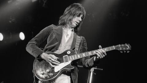 A black and white image of Jeff Beck, who is wearing a blazer and has long, dark hair, playing a guitar.